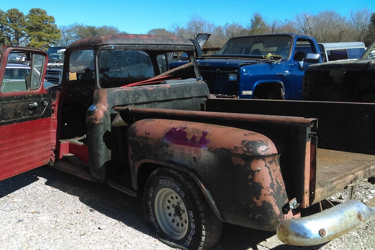 Tonys Auto Salvage: Rusty old pickup with open door in a gravel salvage yard with rows of cars and trucks behind it under a clear blue sky