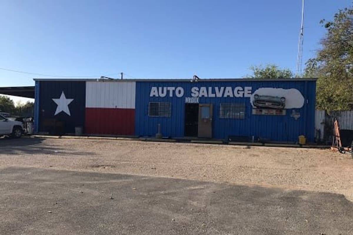 B & T Used Auto Parts: Blue metal auto salvage building painted with a Texas flag, with a gravel lot in front under a clear sky