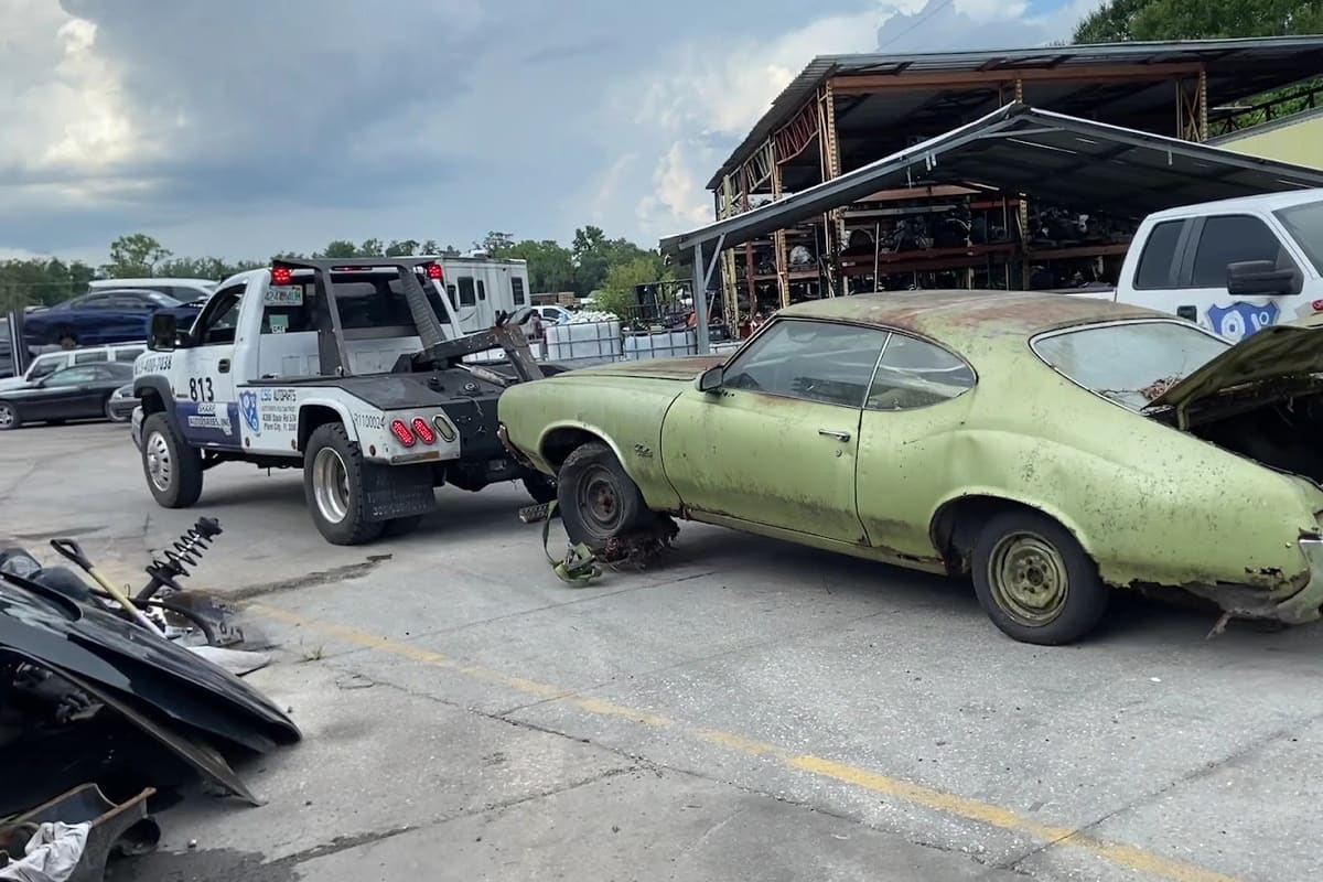CSG Auto Parts: Tow truck pulling a rusty green classic coupe across a salvage lot with covered parts racks and other vehicles behind