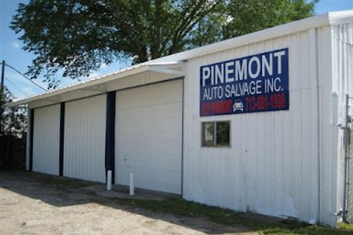 Pinemont Auto Salvage: White metal shop building with large roll-up doors and a blue wall sign, beside a gravel lot under a tree