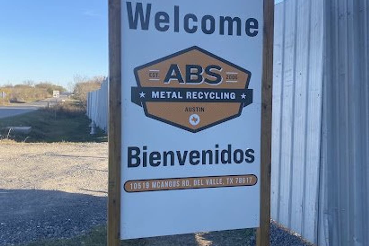 ABS Metal Recycling: Welcome sign in English and Spanish beside a corrugated metal fence and gravel drive under a clear blue sky