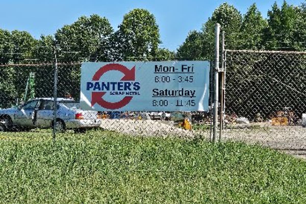 Panter's Garage and Auto Salvage: Hours banner on a chain-link fence beside a gravel lot with a parked car and scrap piles behind it