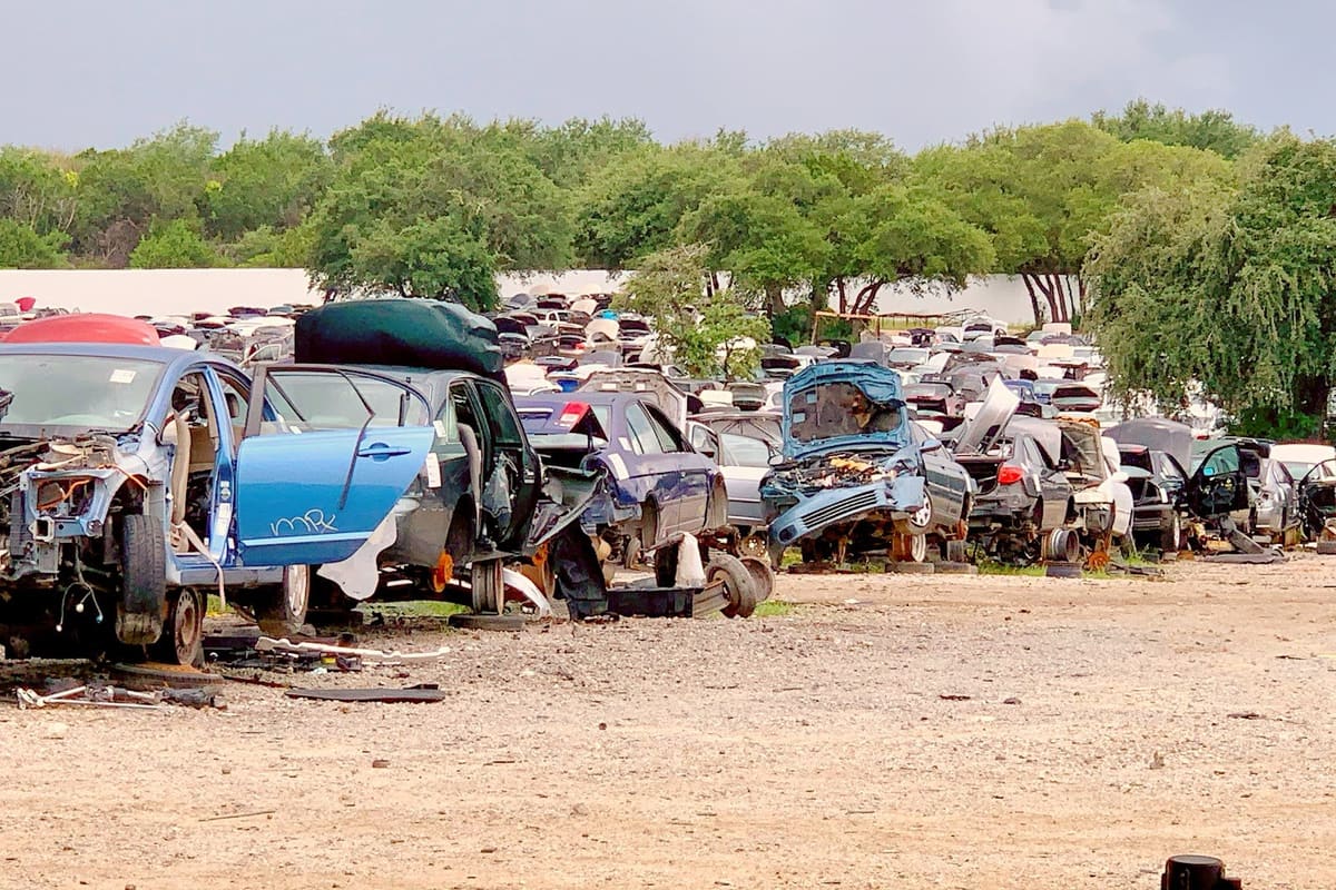 Pick Your Part - Austin: Wide view of a dirt salvage yard with rows of wrecked cars under a cloudy sky and trees in the background