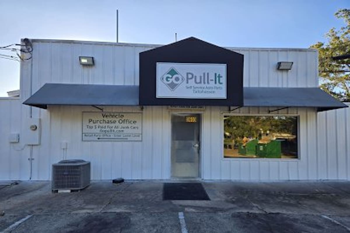 Pick-n-Pull: White metal office building with a black gable sign over the front door and a parking area in front