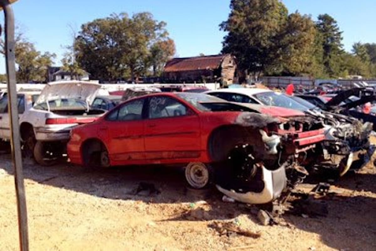 Auto Parts U Pull - Shelby: Rows of wrecked cars parked on a dirt lot under a clear blue sky, with trees and an old barn in the background