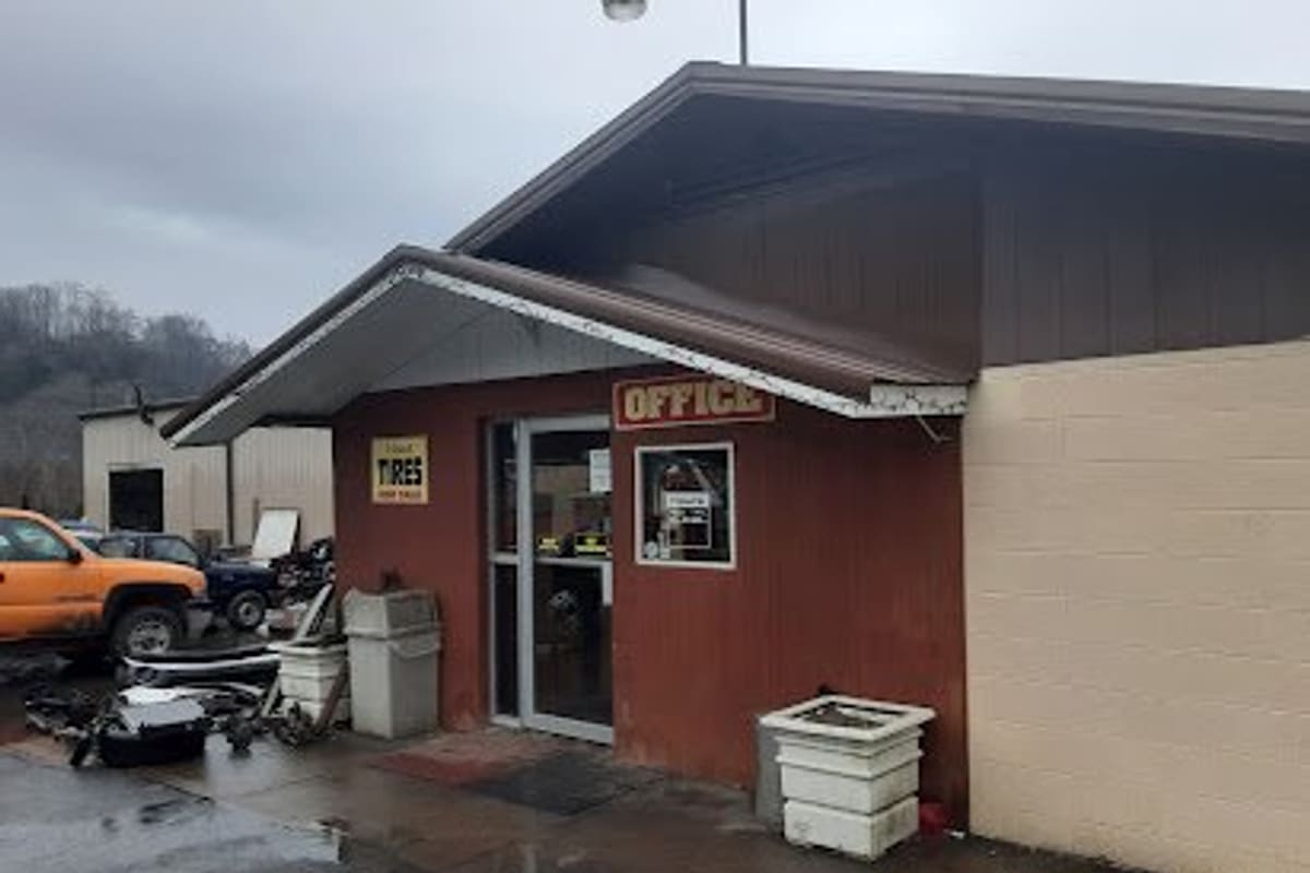 Blountville Auto Salvage: Red-brown office building with a glass door and an Office sign, wet pavement and parts in front, and a truck nearby
