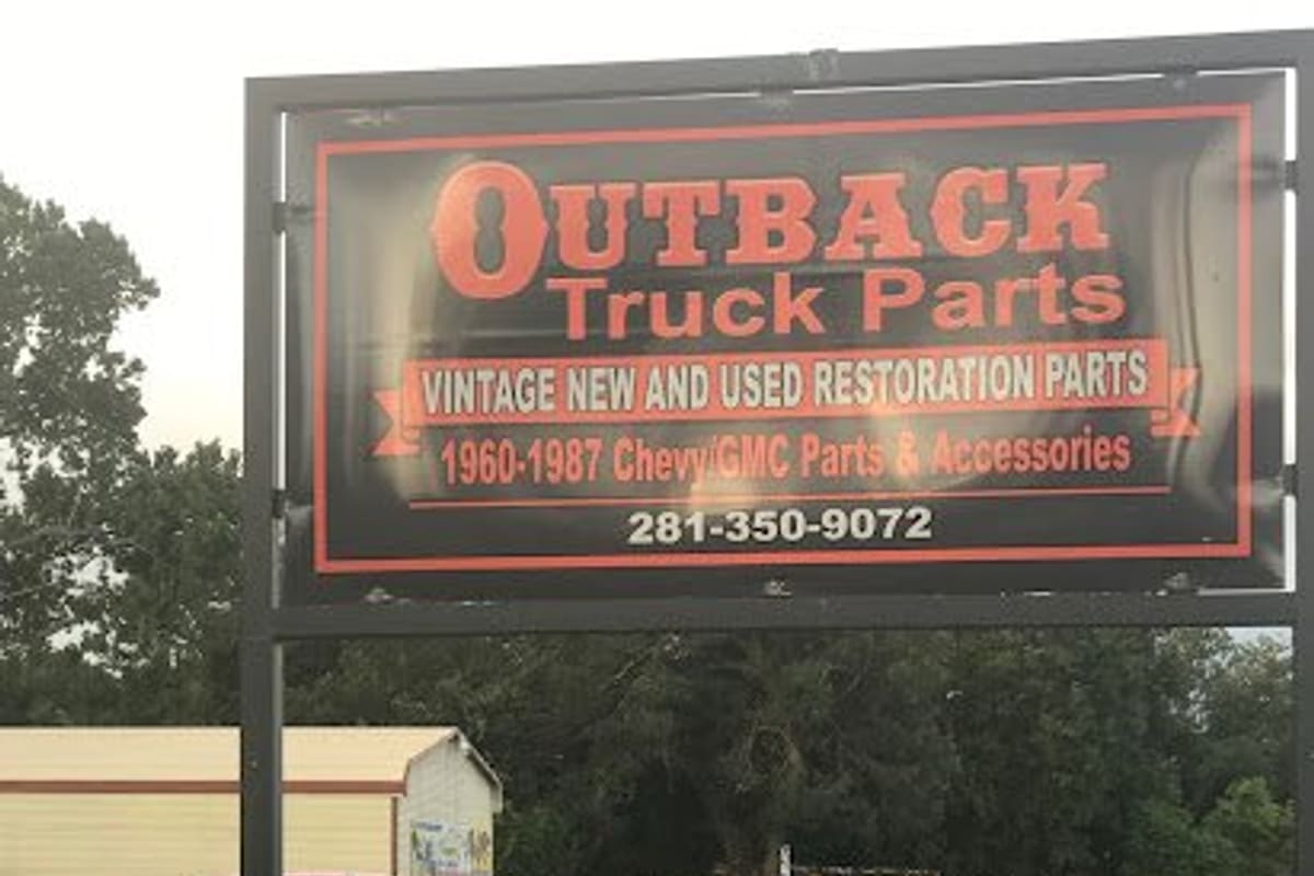 Outback Truck Parts: Large black and orange business sign beside a grassy lot with vintage pickup trucks parked in the background