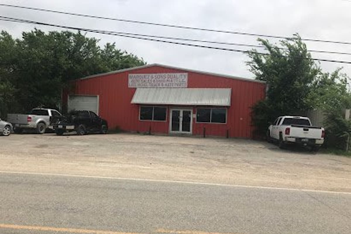 Marquez & Sons Quality Auto & Used Parts: Red metal shop building with a sign above the awning, glass double doors, and pickup trucks parked on a gravel lot beside the road