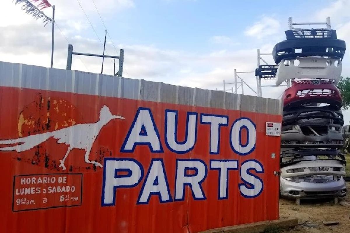 El Paisano Used Auto Parts: Red corrugated metal wall painted with a roadrunner and the words Auto Parts, with stacked car bumpers beside it