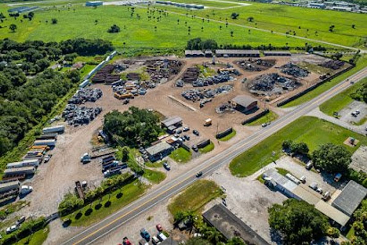 Atland Recycling: Aerial view of a large salvage yard with piles of scrap metal and vehicles beside a highway, surrounded by green fields