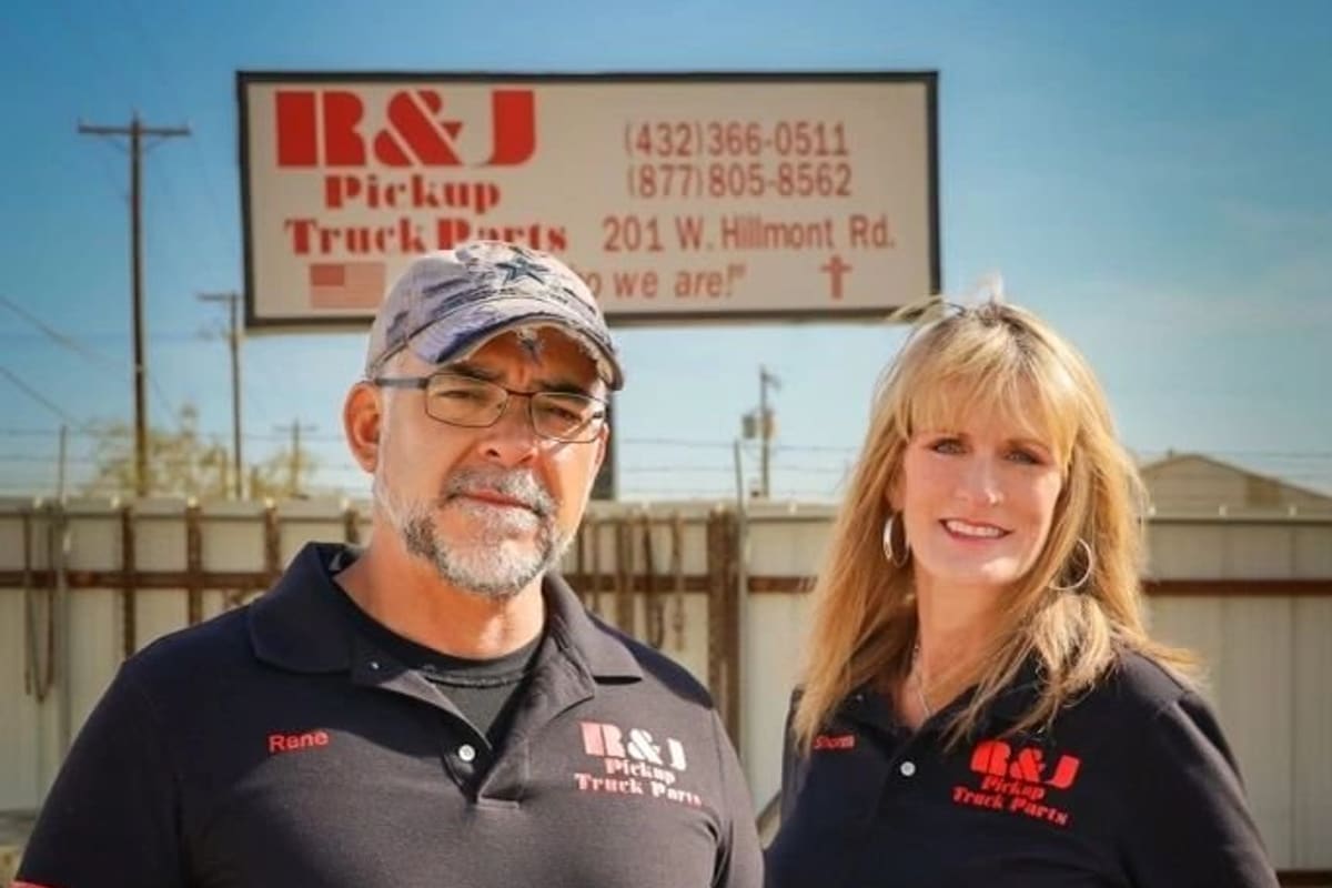 R&J Truck Parts: Man and woman in black work shirts standing in a dirt yard in front of a roadside sign with phone numbers and street address