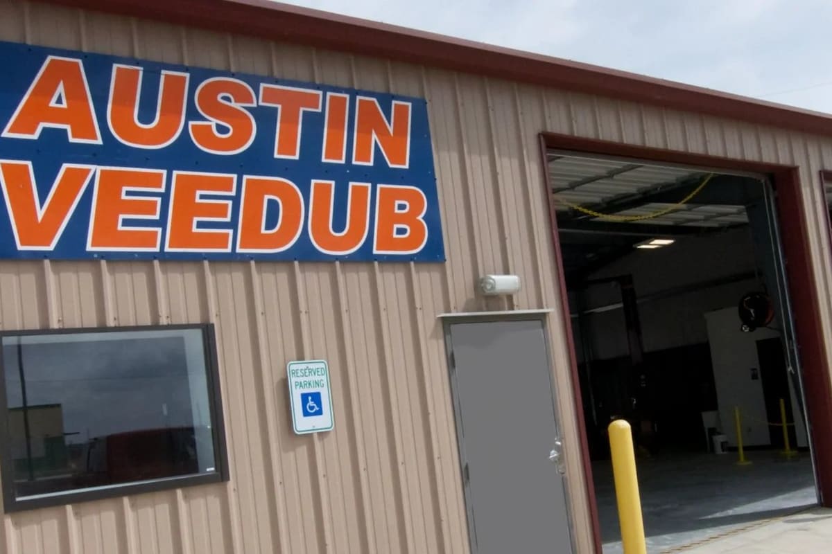 Austin Vee Dub: Tan metal shop building with a large blue and orange sign, a gray door, and open garage bays with yellow posts