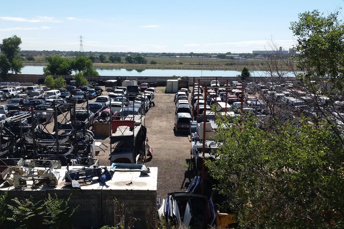 Jim's Used Toyota Parts: Elevated view of a salvage yard with rows of cars and stacked parts, with trees and a lake in the background