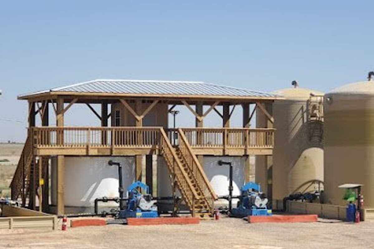 Pecos Yard: Elevated wooden platform with stairs over blue pumps, beside large tan storage tanks on a dirt lot under clear sky
