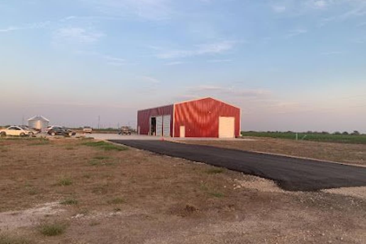 Toms Auto Service: Red metal shop building with roll-up doors beside a fresh asphalt driveway on open flat land, with cars parked nearby