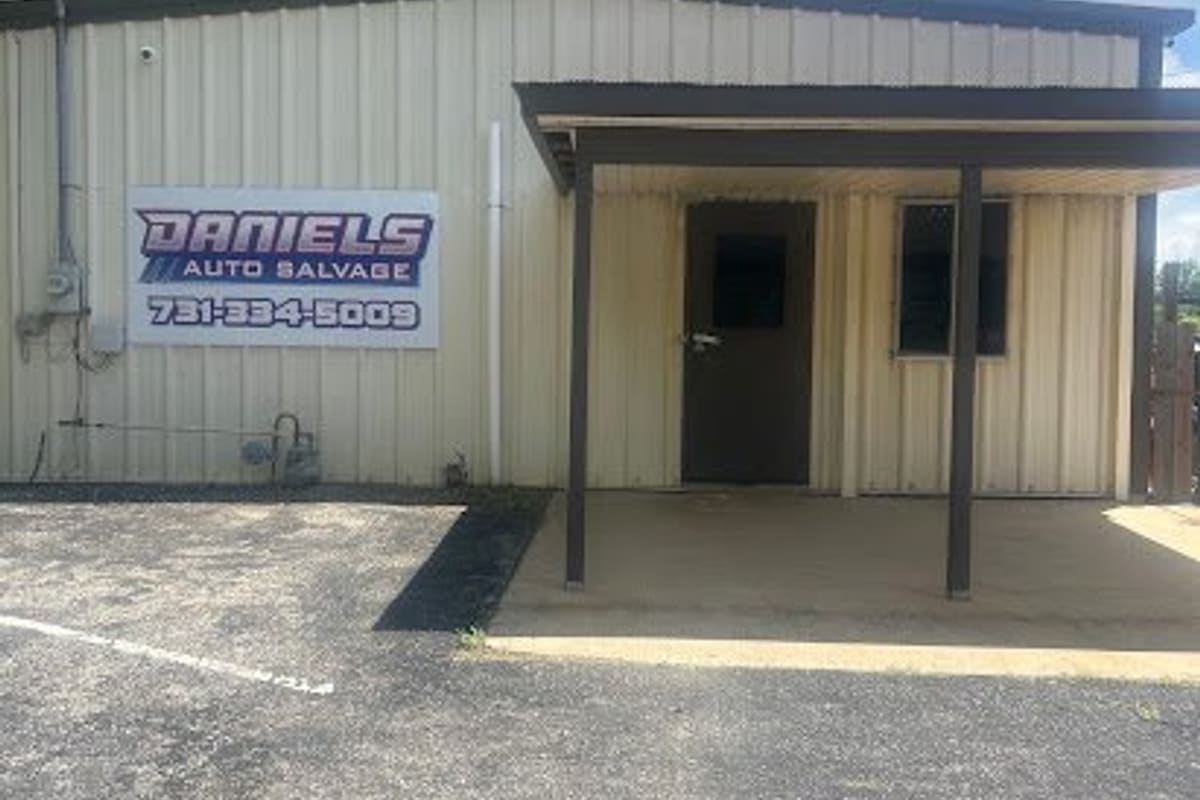 Daniels Auto Salvage: Tan metal office building with a covered entrance, a wall sign with a phone number, and a paved gravel parking area in front