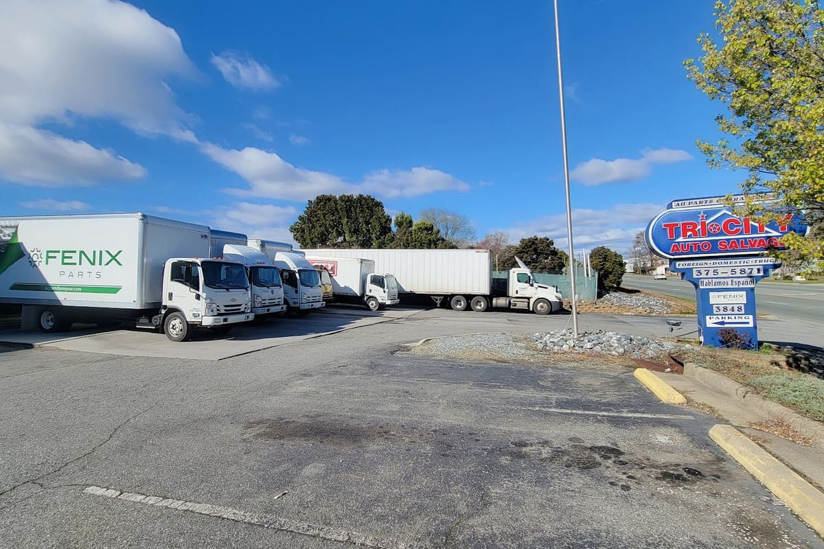 Fenix Parts-Greensboro: Roadside auto salvage sign beside a parking lot with white delivery box trucks parked under a blue sky
