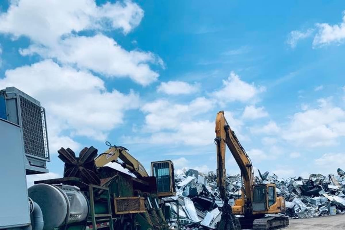 Wilco Iron and Metal Recycling: Excavator and green shredder equipment beside large piles of scrap metal in a gravel recycling yard under a blue sky