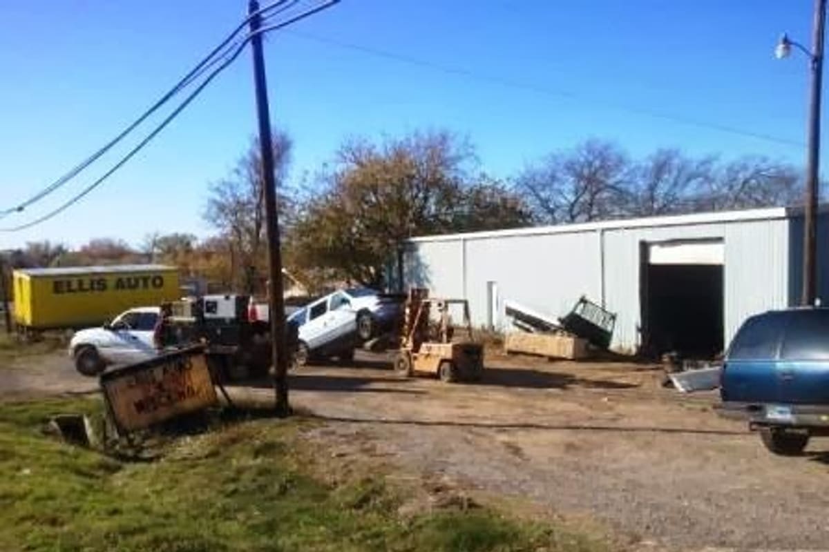 Ellis Auto Salvage: Gravel salvage yard with a forklift, cars, a metal warehouse building and a yellow truck under a clear blue sky