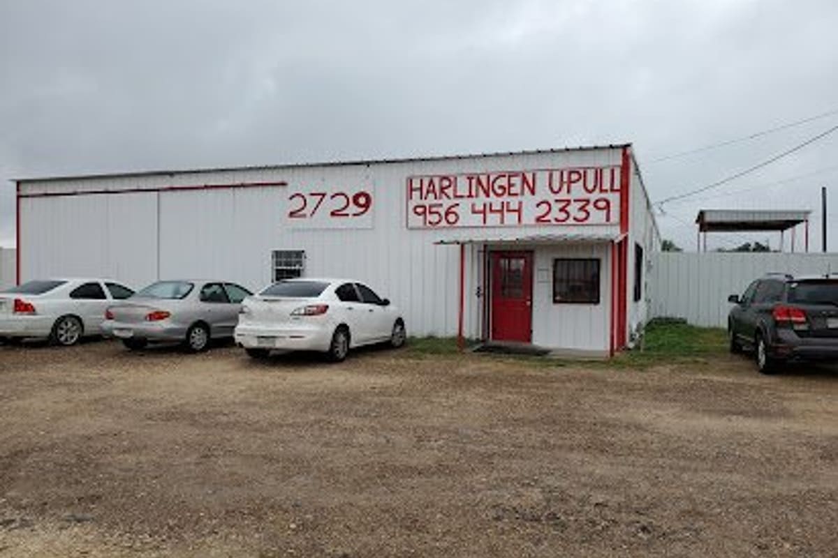 Harlingen U-Pull: White metal building with a red door, painted sign and phone number, and cars parked on a gravel lot under a cloudy sky