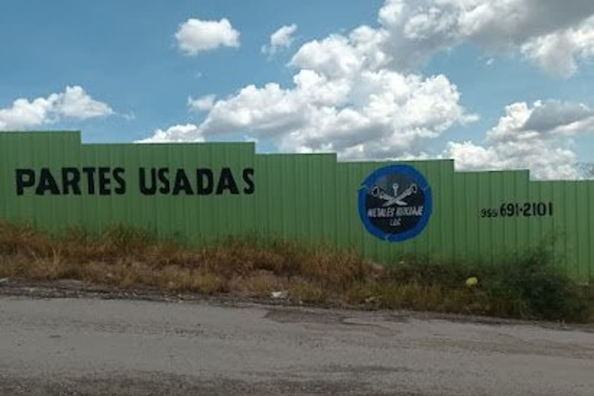Metales Reciclaje: Green metal fence painted with the words Partes Usadas, a round logo and a phone number, beside a gravel road