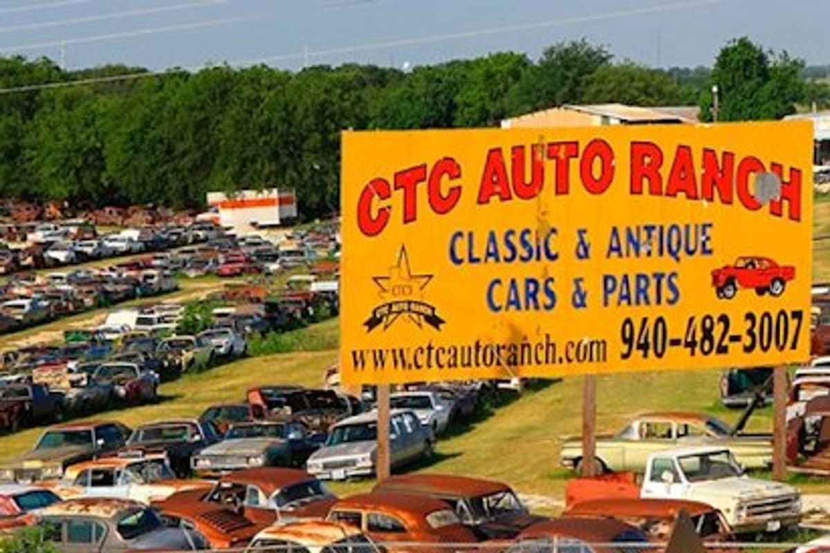 CTC Auto Ranch: Large yellow roadside sign advertising classic and antique cars and parts above rows of rusty vehicles in a salvage yard