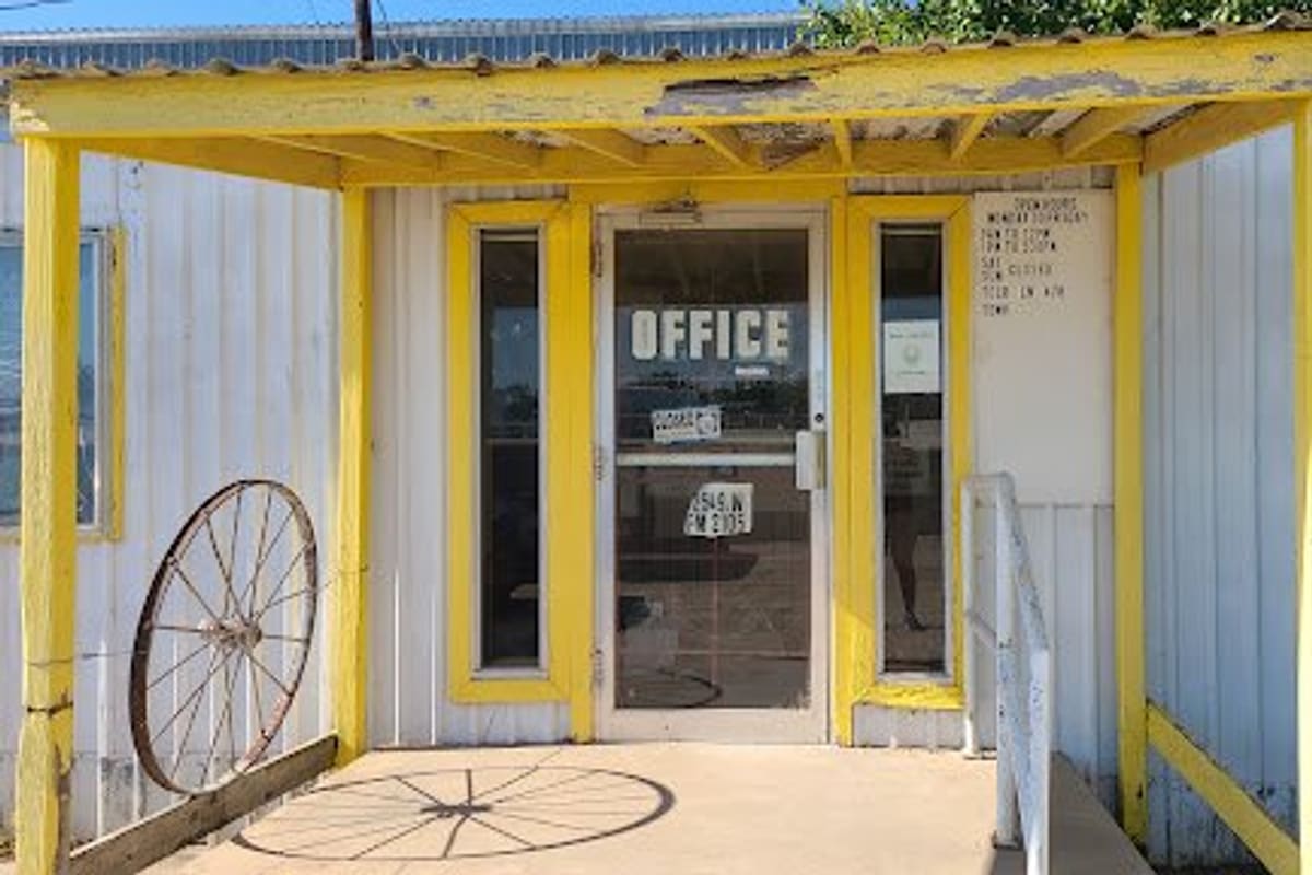 Neiras Wrecking Yard: Yellow-trimmed covered entrance to a small office with glass door, hours posted beside it, and a wagon wheel on the left