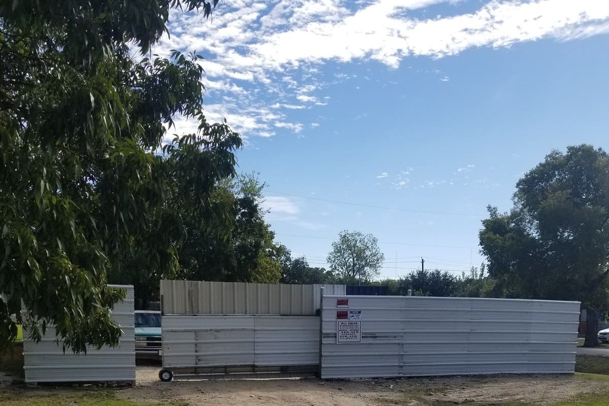 South Texas Auto Sales and Salvage: Metal-paneled fence and rolling gate at the end of a gravel drive, seen from a cracked asphalt road on a sunny day