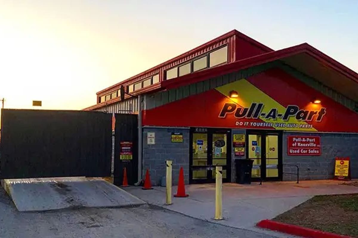 Pull-A-Part Knoxville: Red and yellow self-service auto parts building with glass entrance doors, gated fence and orange cones at dusk