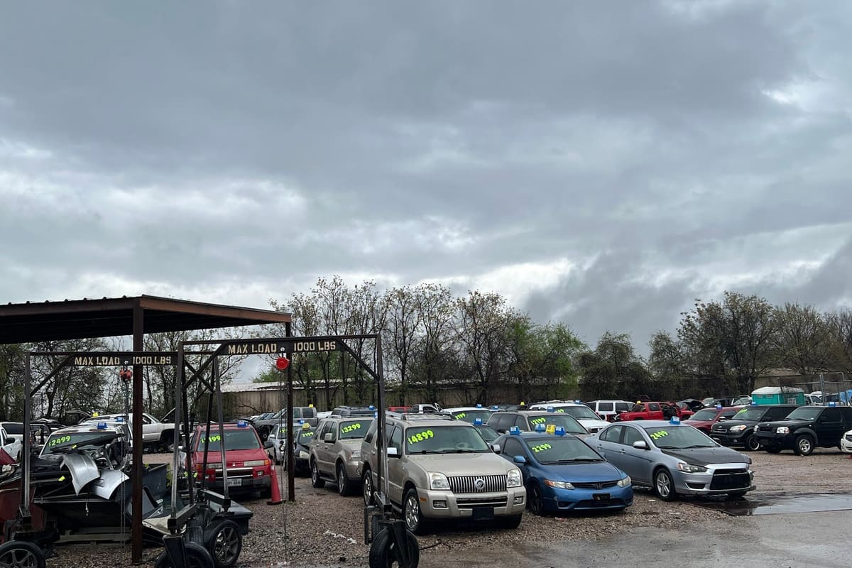 Pick-n-Pull - Fort Worth: Wide view of a gravel lot with rows of parked cars with price tags under a cloudy sky