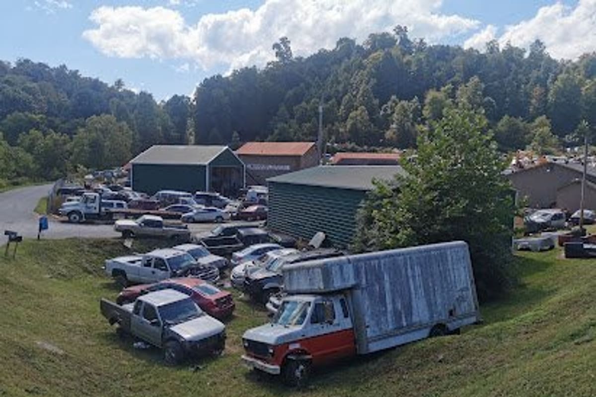 Roan Mountain Auto Salvage: Elevated view of a salvage yard with trucks and cars parked on a grassy slope in front of green metal sheds and wooded hills