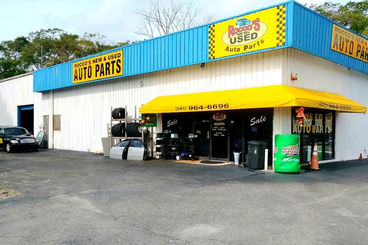 Rocco's Auto Parts: Auto parts store with blue and white metal siding, yellow awning, tire rack and glass entrance doors on an asphalt lot