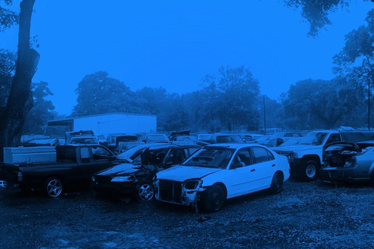 Angelos Auto Parts: Rows of wrecked and used cars parked on a gravel lot beneath tall trees, with a white building in the background