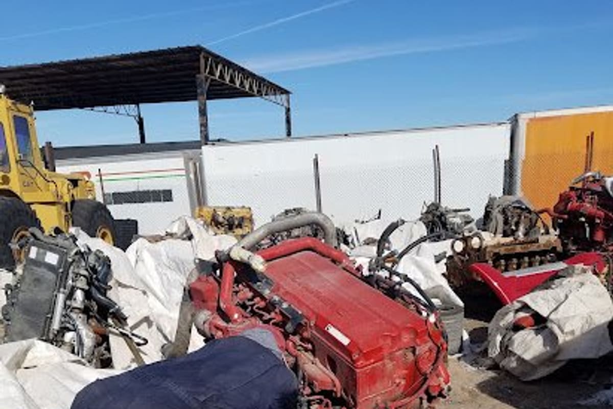 Pardo Truck Center: Salvage yard with truck engines and parts piled on tarps, heavy equipment and trailers in the background under a blue sky