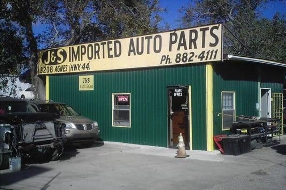 J & S Imported Auto Parts: Green metal auto parts building with a yellow sign, parts office door and vehicles parked in front of a paved lot