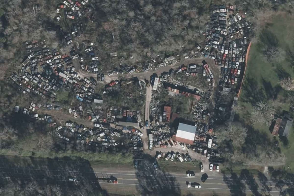 Bradford Auto Parts & Salvage: Aerial view of a salvage yard with rows of vehicles among trees, a warehouse building and a road along the bottom