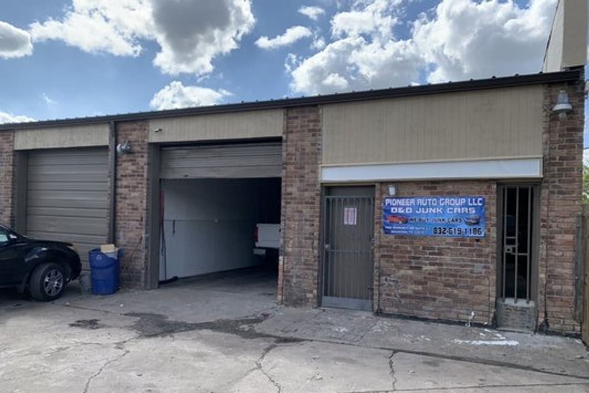 D&D Junk Cars: Brick commercial building with two roll-up garage doors, a metal gate entrance and a banner sign above a cracked concrete lot