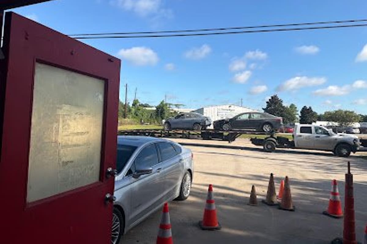 Premier Salvage: Open red door looking out onto a gravel lot with traffic cones, a parked sedan and a truck hauling cars on a trailer