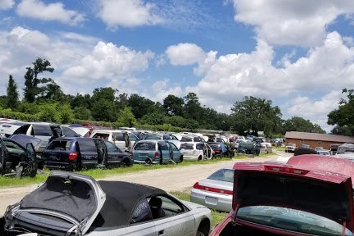 Ocala Junk & Auto Parts: Rows of salvage cars parked on a gravel lot under a blue sky with clouds, with low buildings in the distance