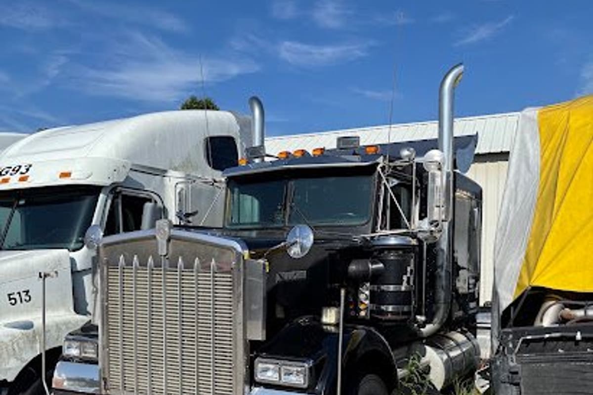 C & H Truck & Equipment: Black semi truck tractor parked on grass and dirt beside other trucks in a salvage yard under a blue sky