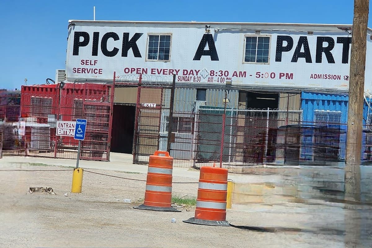 RG Pick-A-Part: Two-story white building with a large PICK A PART sign, fenced with razor wire, and orange barrels near the entrance