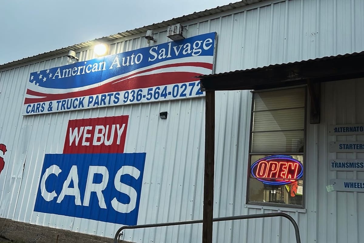 American Auto Salvage: White metal building with a large painted flag sign, a We Buy Cars mural, a lit open sign and a blue entrance door