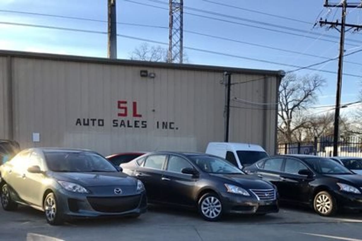 SL Auto Sales: Row of parked used cars in front of a tan metal building with a painted business sign and a fence behind