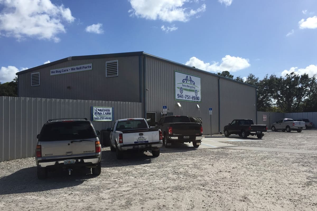 A & a Auto Recycling: Gray metal warehouse building with signs reading We Buy Cars, We Sell Parts and Cash for Junk Cars, trucks parked on a gravel lot