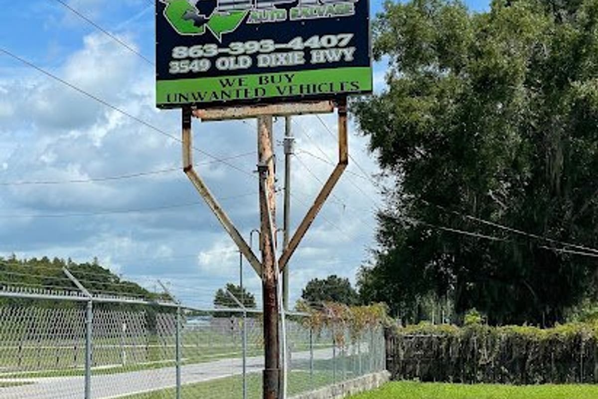 Epic Auto Salvage: Tall roadside sign on a rusty metal pole beside a grassy lot, advertising a phone number, street address and vehicle buying