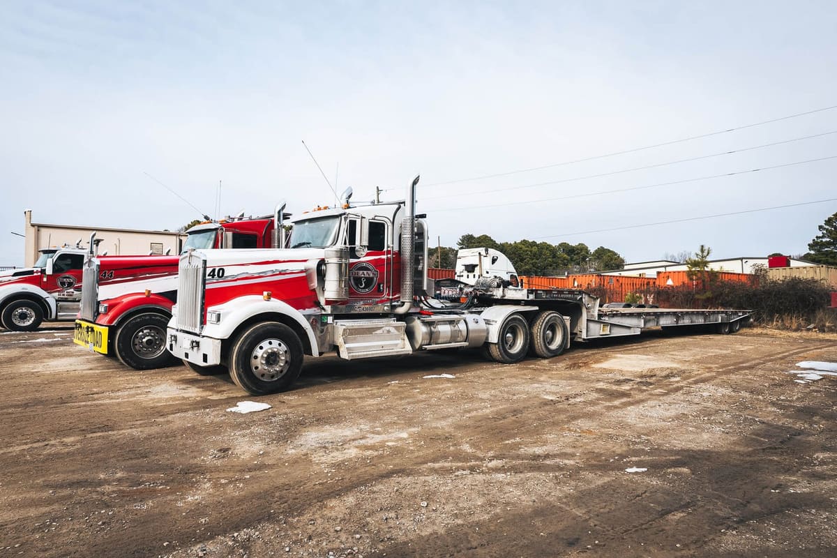 Dean's Wrecker Service: Red and white heavy-duty tow trucks and a lowboy trailer parked on a dirt lot beside industrial buildings