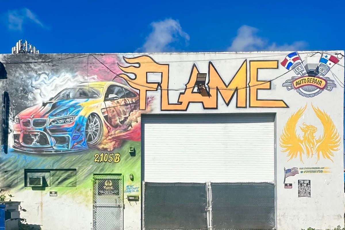 Flame Parts: White warehouse-style building with a large colorful car mural, orange flame lettering, and a roll-up garage door above an empty parking lot