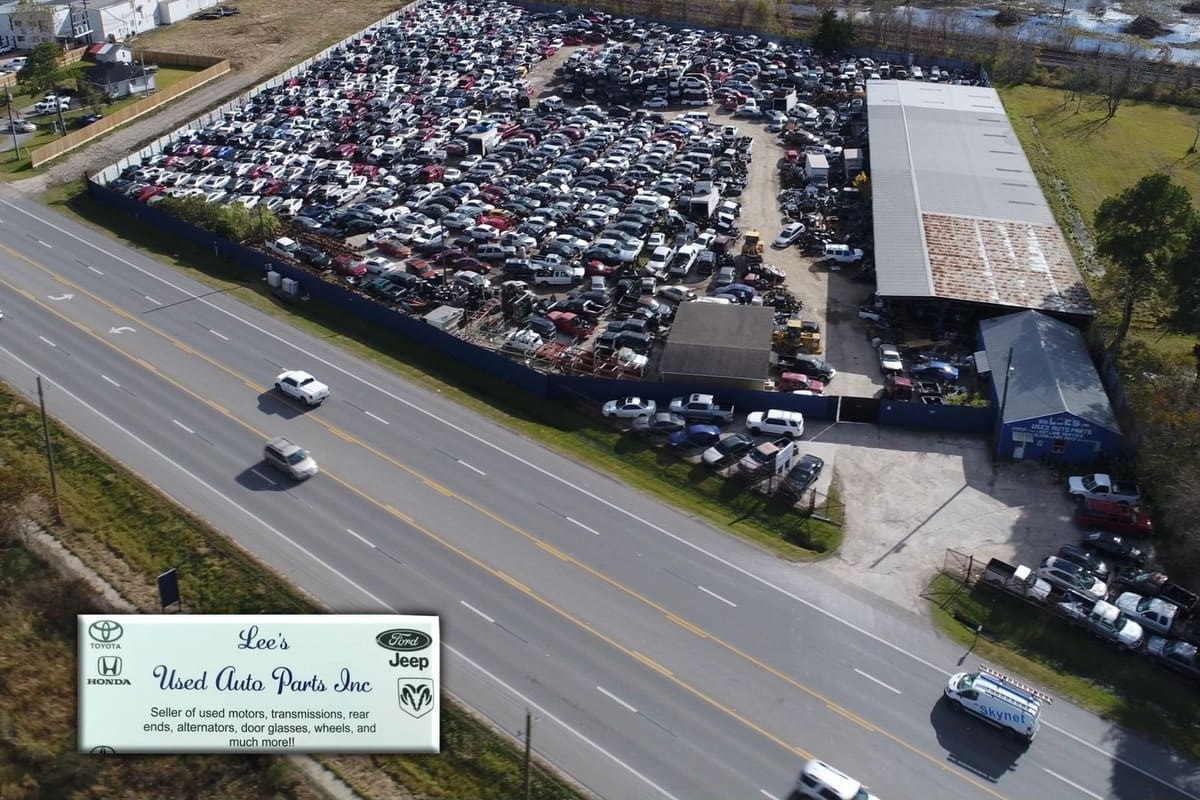Lee's Used Auto Parts: Aerial view of a large salvage yard with rows of vehicles beside a highway, with warehouse buildings and a blue office building