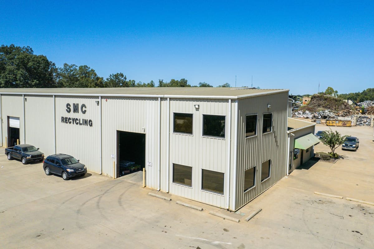 SMC Recycling: Aerial view of a two-story metal industrial building with recycling sign, parked vehicles, and scrap piles in the background