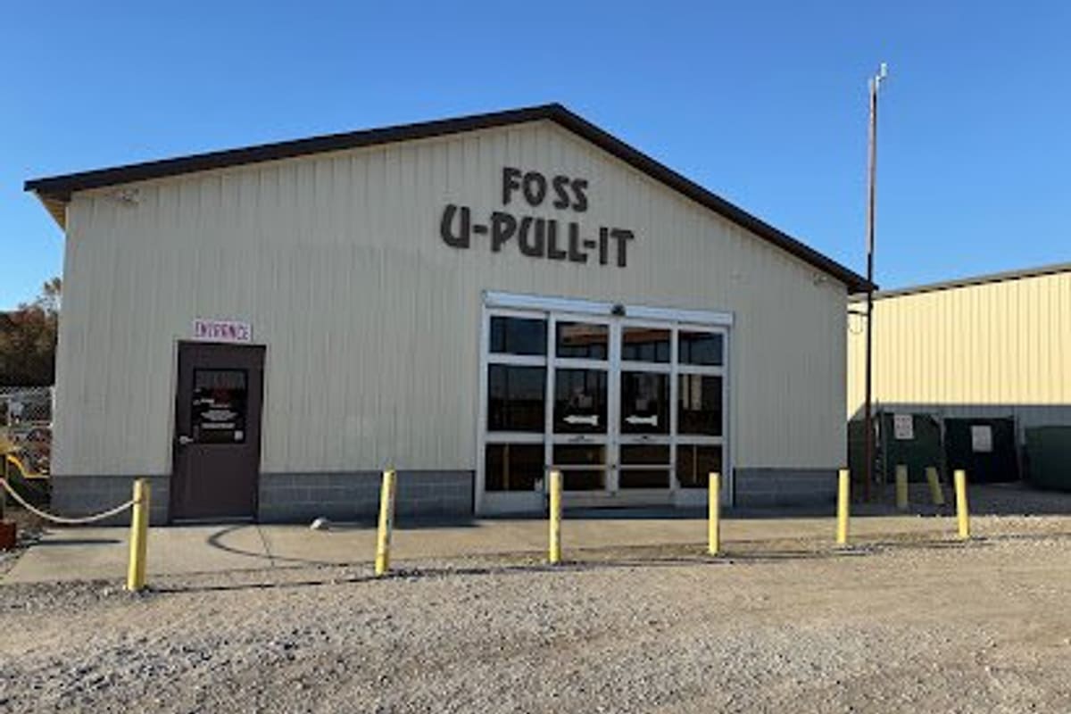 Foss U-Pull-It La Grange: Cream metal building with large lettering on the front, a side entrance door, glass sliding doors, and yellow posts over a gravel lot